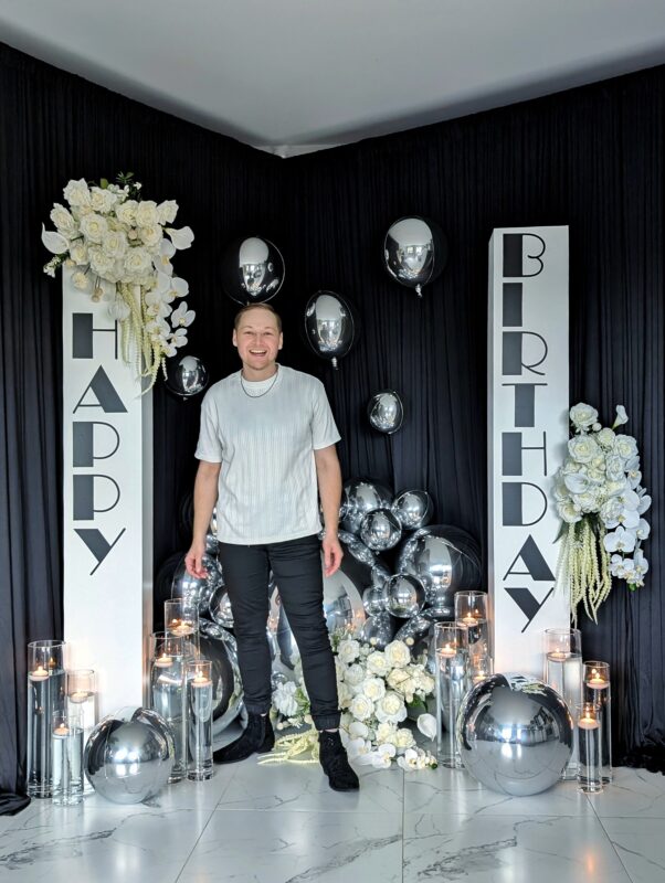 Pawel standing in front of a birthday display with silver mirror ball balloons, candles and white florals.