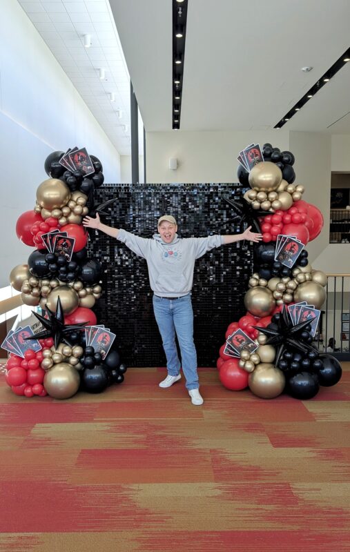 Pawel Rzeznik standing with a polished black, red and gold balloon backdrop.