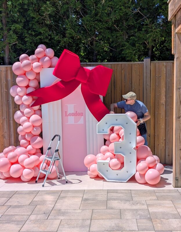 Pawel setting up a third birthday party with pink balloons, a huge red bow and a marquee number three.