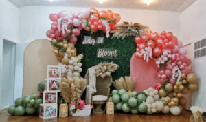 Baby shower event decor featuring two arch walls, boxwood backdrop, wicker chair, and balloons.