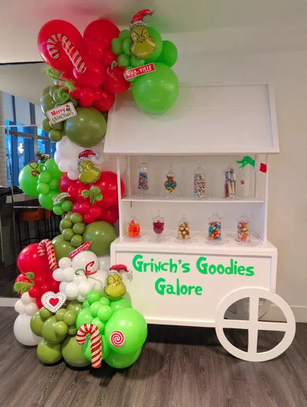 Festive balloon display featuring the Grinch and candy-themed decor around a sweets cart labeled "Grinch's Goodies Galore."