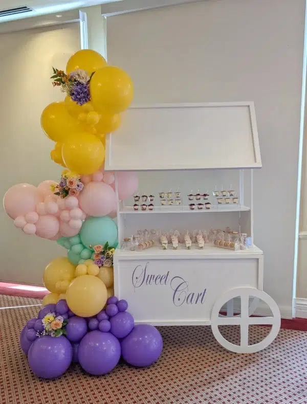 A white sweet cart adorned with colorful balloons and filled with desserts, perfect for celebrations or events.