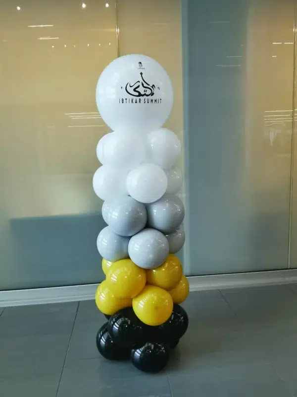 A decorative balloon tower featuring white, gray, yellow, and black balloons, topped with a logo for Ibtikar Summit.