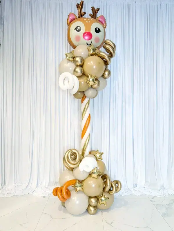 A whimsical balloon arrangement featuring a cute reindeer head, gold and cream balloons, and playful spirals, set against a white backdrop.