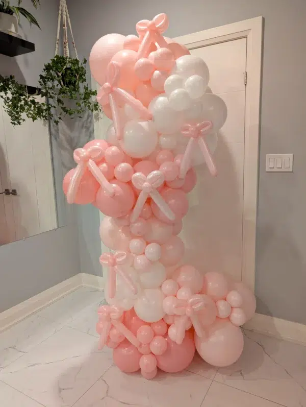 A vibrant balloon arrangement featuring pink and white balloons, adorned with ribbon bows, set against a pastel wall.