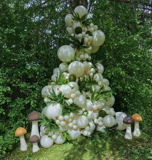 A whimsical display of white balloons arranged in a pyramid shape, accented by greenery and decorative mushrooms in a forest setting.