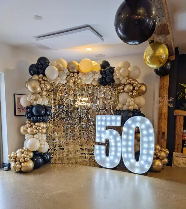 A festive birthday setup featuring balloons in black, gold, and cream, a sequined backdrop, and illuminated "50" sign.