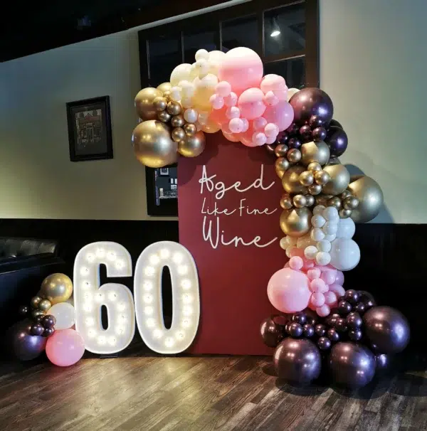 A festive balloon arch featuring shades of pink, gold, and purple with a light-up "60" and the phrase "Aged Like Fine Wine."