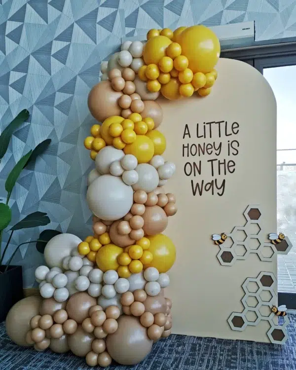 Elegant balloon display featuring gold, white, and cream colors with a sign saying "Our Little One Is On The Way," set in a modern room.