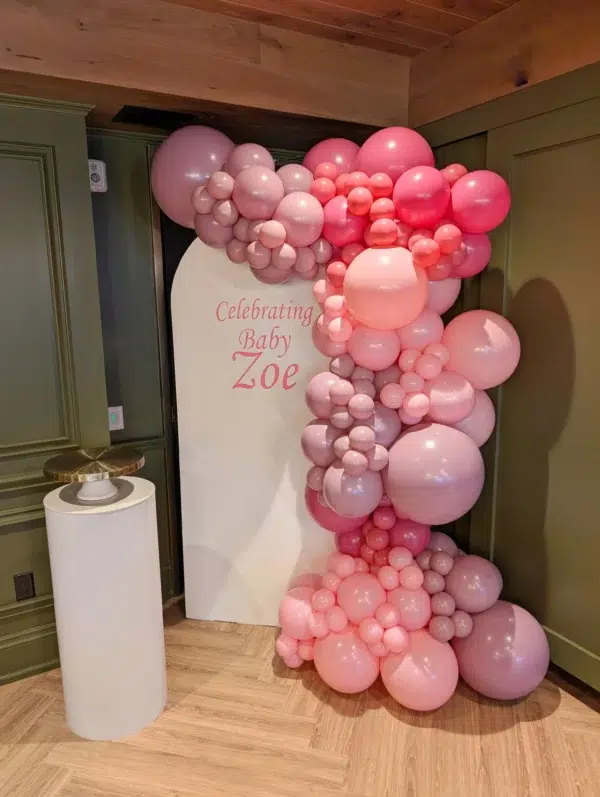 Colorful balloon arch in shades of pink celebrating "Baby Zoe," with a sleek, white pedestal nearby in a stylish indoor setting.