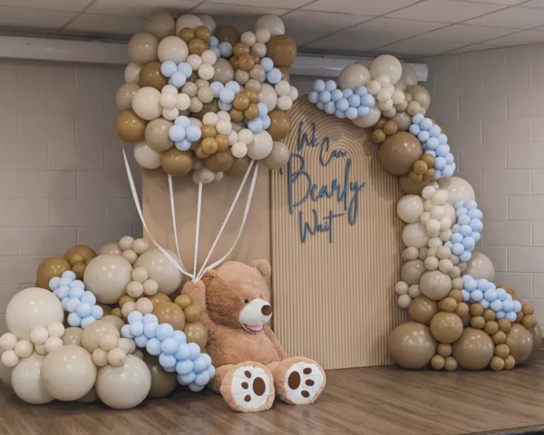 A playful balloon decoration featuring beige and blue balloons, with a large teddy bear sitting beside a backdrop that reads "We Can Bearly Wait."