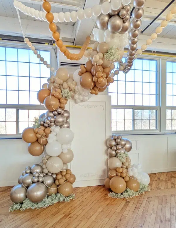 Elegant balloon arch with beige, white, and metallic balloons, adorned with delicate flowers, perfect for celebrations or events.