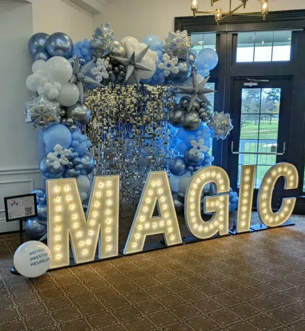 A vibrant event backdrop featuring "MAGIC" in illuminated letters surrounded by blue and silver balloons and decorative elements.