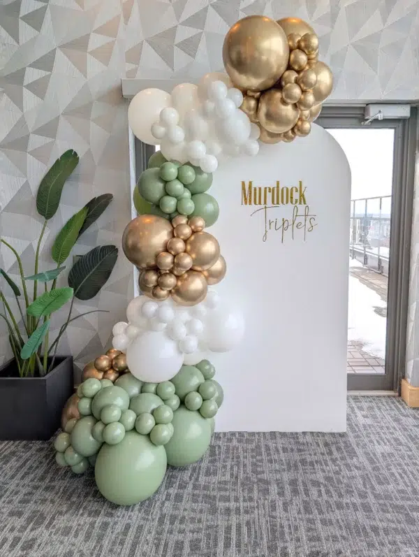 A stylish balloon arch featuring gold, white, and green balloons, with a sign reading "Murdock Triplets" in a modern setting.