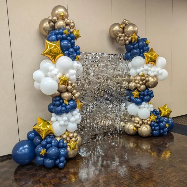 Colorful balloon arch with gold, white, blue, and star accents in front of a shiny mirrored backdrop, set in an indoor venue.