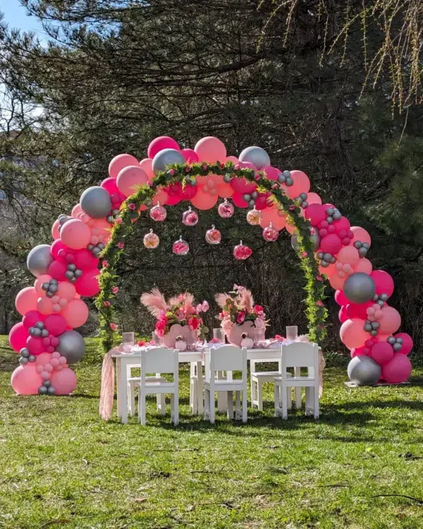 Colorful balloon arch in pink, silver, and gray, framing a festive outdoor dining setup with floral centerpieces and white chairs.