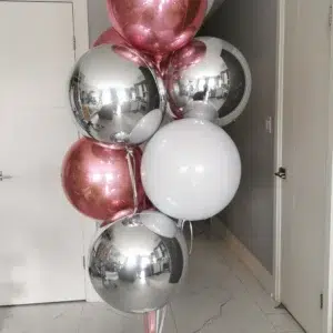 A cluster of decorative balloons in pink, silver, and white, arranged with a white balloon base, set against a light gray wall and door.