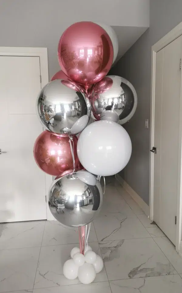 A cluster of decorative balloons in pink, silver, and white, arranged with a white balloon base, set against a light gray wall and door.