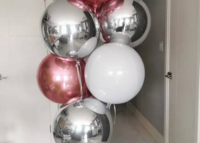 A cluster of decorative balloons in pink, silver, and white, arranged with a white balloon base, set against a light gray wall and door.