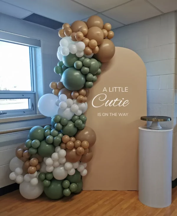 Decorative balloon display in earthy tones with a sign reading "A Little Cutie Is On The Way," set against a neutral backdrop.