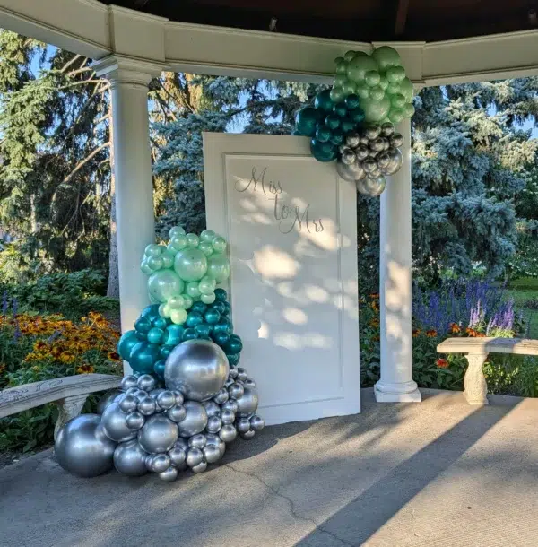 A decorative arch made of teal, green, and silver balloons with a white door featuring the text "Miss to Mrs. in a garden setting.