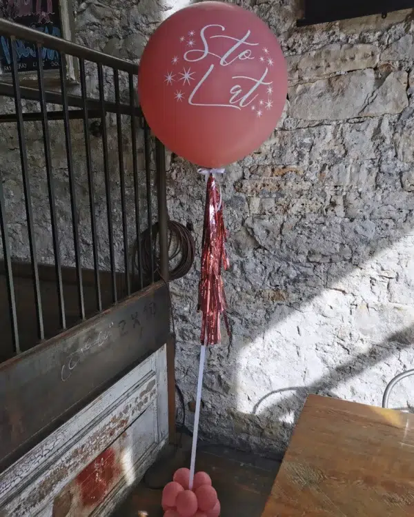 A large, rose-colored balloon with white lettering and tinsel accents stands on a decorated base, against a stone wall backdrop.