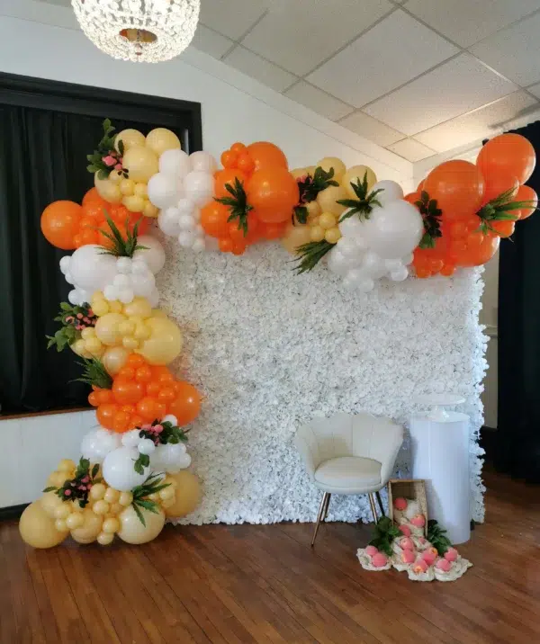 Colorful balloon arch with orange, yellow, and white balloons above a white flower wall and a white chair in a decorated venue.