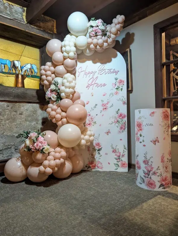 Decorative birthday display with pink balloons, flowers, and a sign saying "Happy Birthday" against a rustic backdrop.