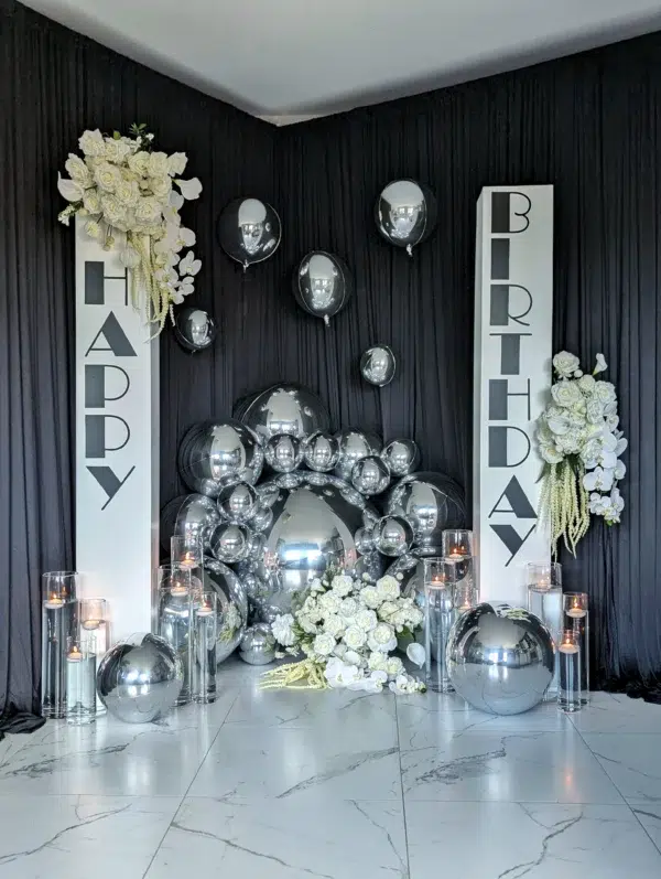 Elegant birthday decor featuring white flowers, metallic balloons, and stylish silver accents against a black backdrop. Perfect for celebrations.