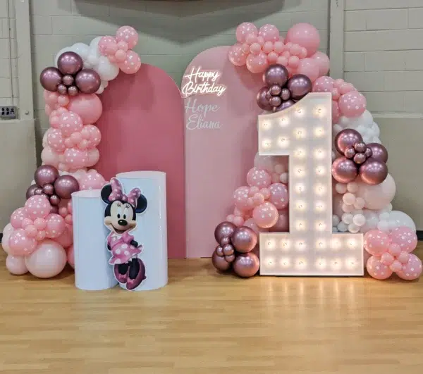 Decorative birthday setup with pink balloons, a large illuminated "1", and a Minnie Mouse cutout for a child's first birthday celebration.