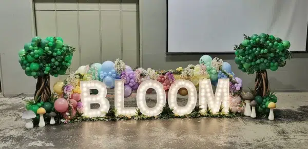 Colorful balloon display featuring the illuminated word "BLOOM," surrounded by floral accents and playful mushroom decorations.