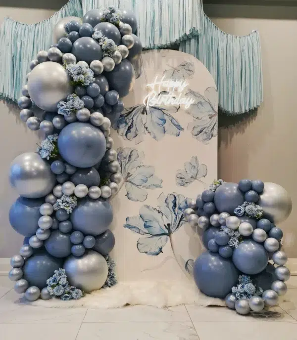 Decorative balloon arch in shades of blue and silver with floral accents, featuring a "Happy Birthday" sign and elegant backdrop.