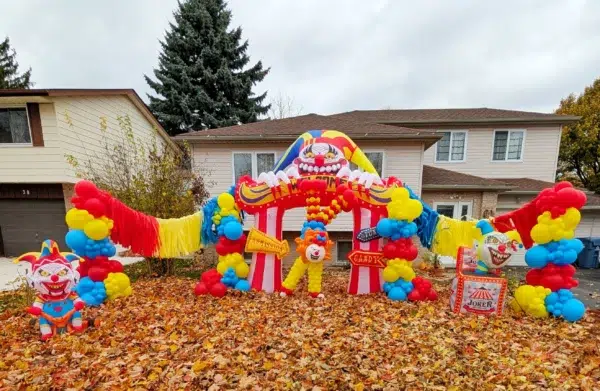 Colorful clown-themed inflatable decoration with balloons, entrance to a spooky carnival, surrounded by autumn leaves in a residential area.