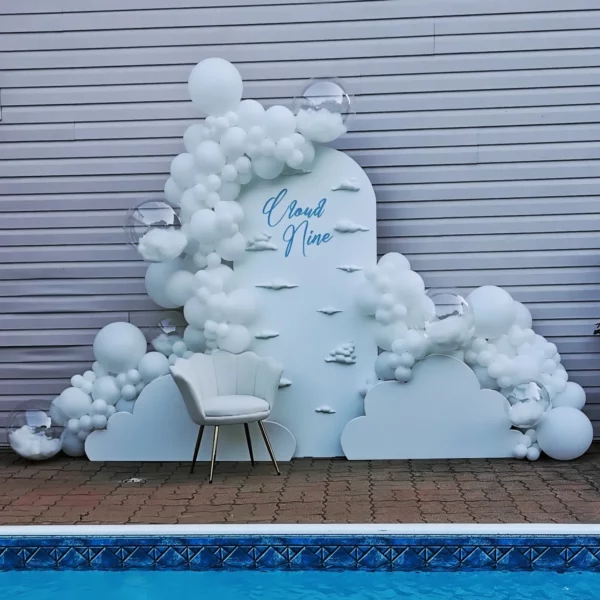 A whimsical backdrop featuring white balloons and clouds reading "Cloud Nine," with a stylish chair in front, set by a pool.