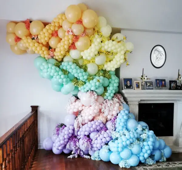 Colorful balloon arrangement featuring pastel shades cascading from a wall to the floor, accented with floral elements. Perfect for celebrations.