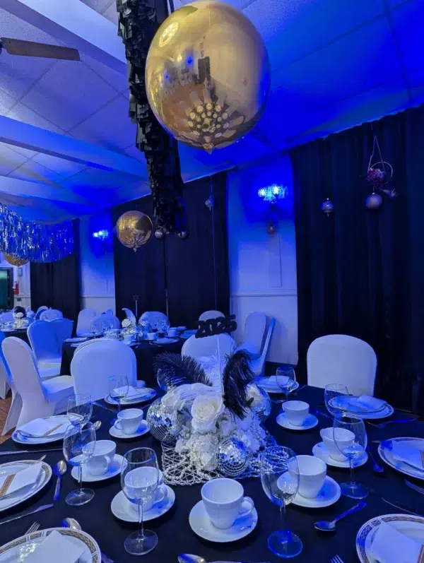 Elegant party setting with blue lighting, golden balloons, and a decorated table featuring floral centerpieces and 2025 signage.