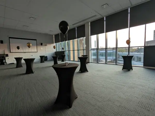 A spacious event room featuring black and gold balloons, cocktail tables, and large windows overlooking an outdoor view.