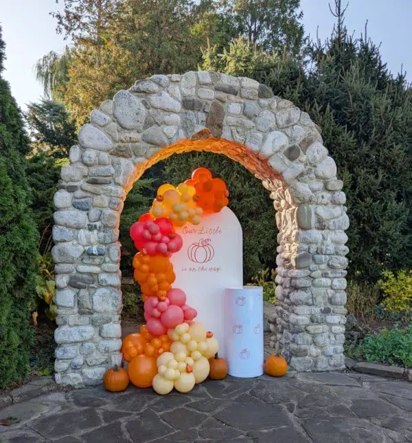 A decorative stone archway with colorful balloons and pumpkins, featuring a sign that reads "Our Little is on the way!" in a festive setting.