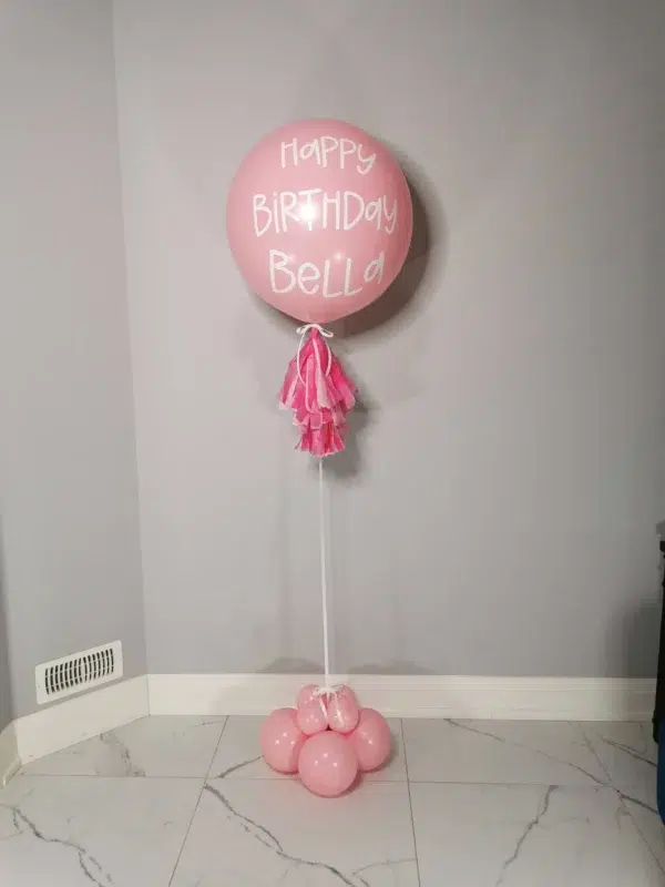 Large pink birthday balloon with the text "Happy Birthday Bella," topped with pink tassels, resting on a cluster of pink balloons.