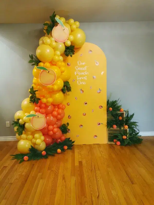 Colorful peach-themed balloon decoration with a yellow backdrop celebrating a first birthday. Green leaves and small peaches enhance the design.