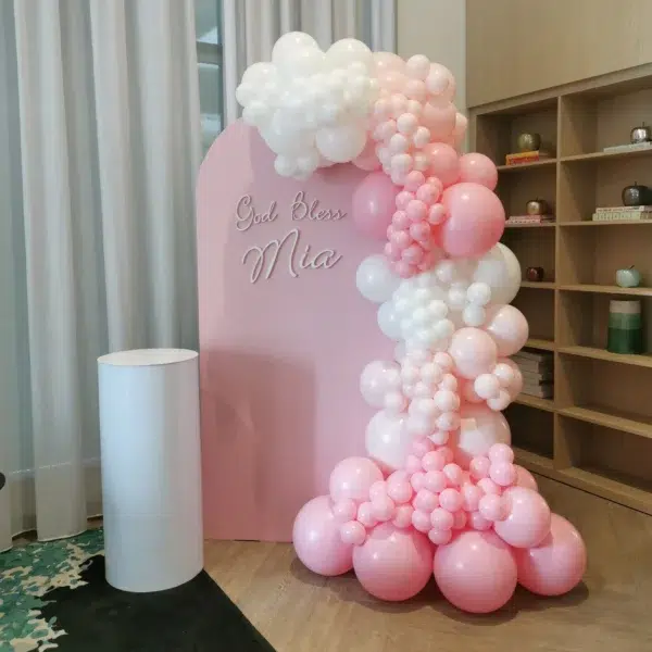 A charming pink balloon arch with the text "God Bless Mia," complemented by a white pedestal and modern decor elements.