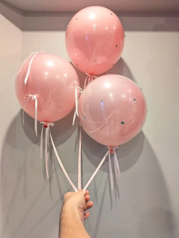 Three pink balloons with decorative embellishments and ribbons, held by a hand against a light gray wall.