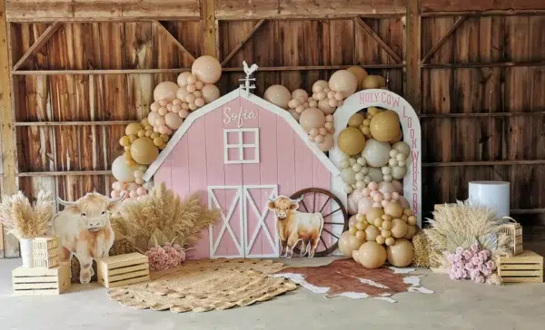 A whimsical barn backdrop featuring pink balloons, a cutout cow, pampas grass, and decorative crates for a festive celebration.