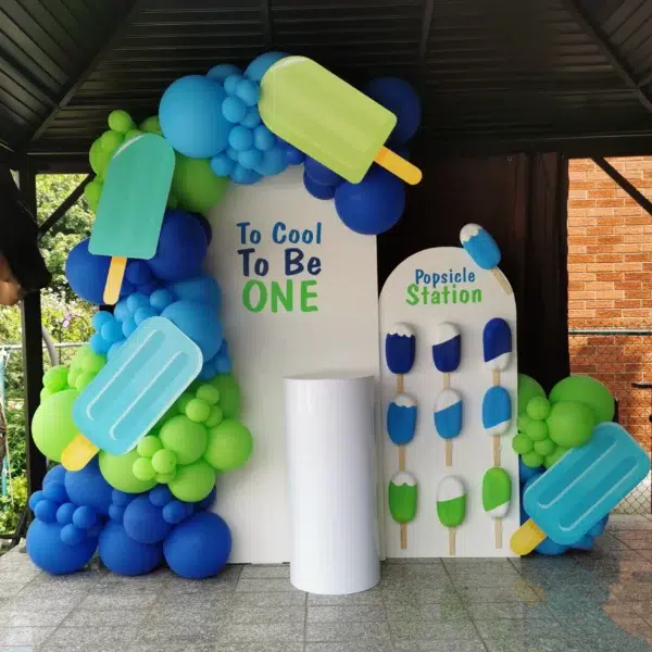 Colorful birthday decoration with popsicle theme, featuring balloons and a sign saying "To Cool To Be ONE" at a popsicle station.