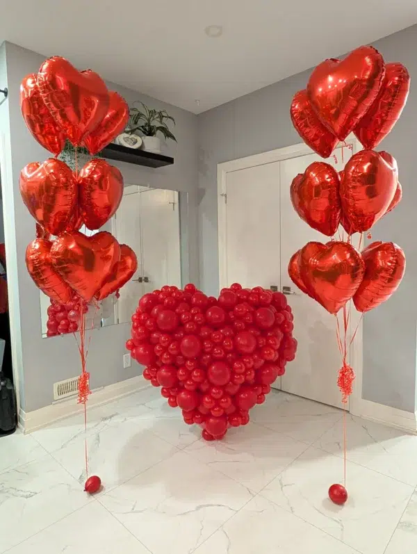 Red heart-shaped balloon arrangement with several heart-shaped balloons on tall strings, set against a light gray wall. Perfect for celebrations.