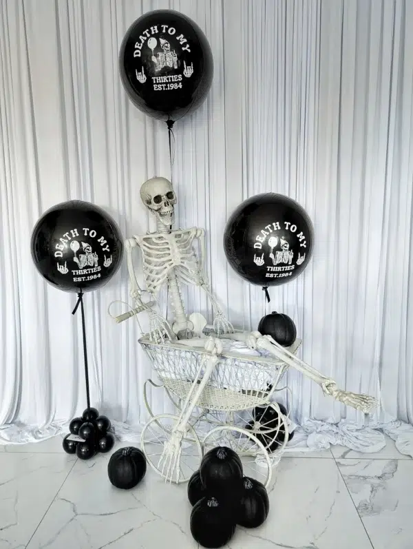 A skeleton sits in a vintage stroller surrounded by black balloons and decorative pumpkins, themed for a humorous 30th birthday celebration.