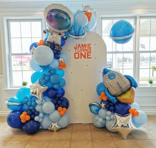 Colorful balloon decor featuring an astronaut and rocket for a first birthday celebration, with "JAMIE TURNS ONE" displayed prominently.