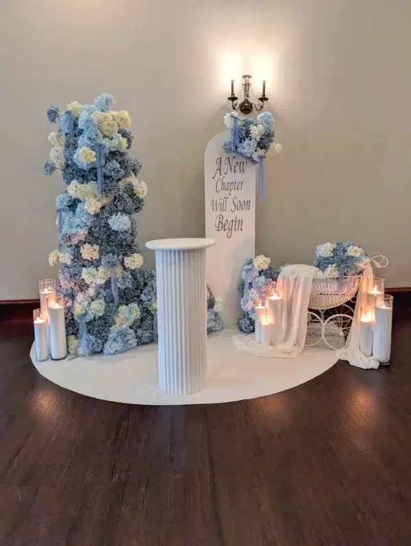 Elegant wedding backdrop with blue and white floral arrangements, a central column, candles, and a sign reading "A New Chapter Will Soon Begin."