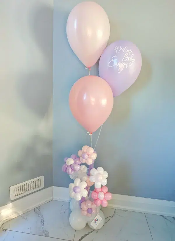 A cluster of pink and purple balloons with a "Welcome Baby" message, accompanied by colorful balloon flowers, against a light wall.