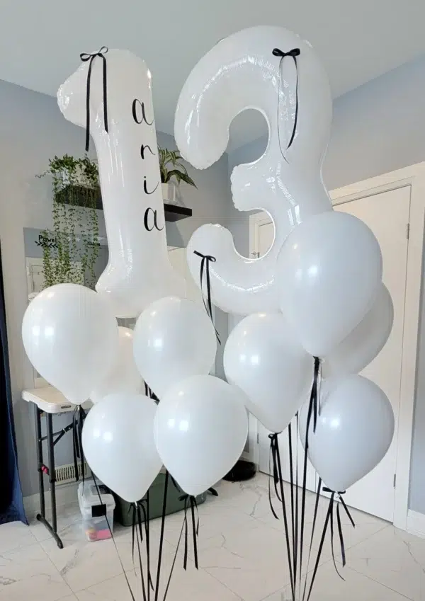 Two large white balloons shaped like "1" and "3" with the name "Aria," surrounded by white balloons with black ribbons.